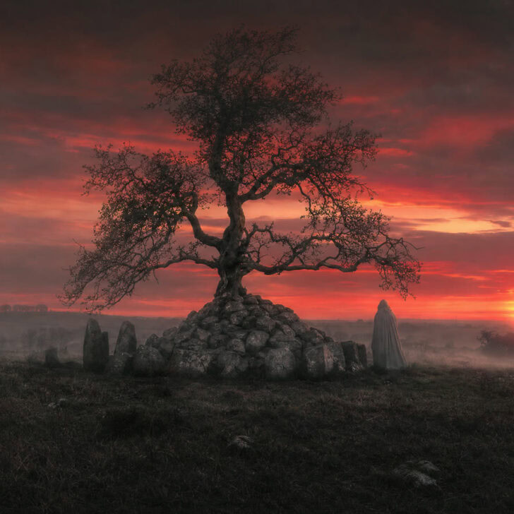 An ancient stone cairn under a hawthorn tree at dusk in County Derry with a faint cloaked figure beside it