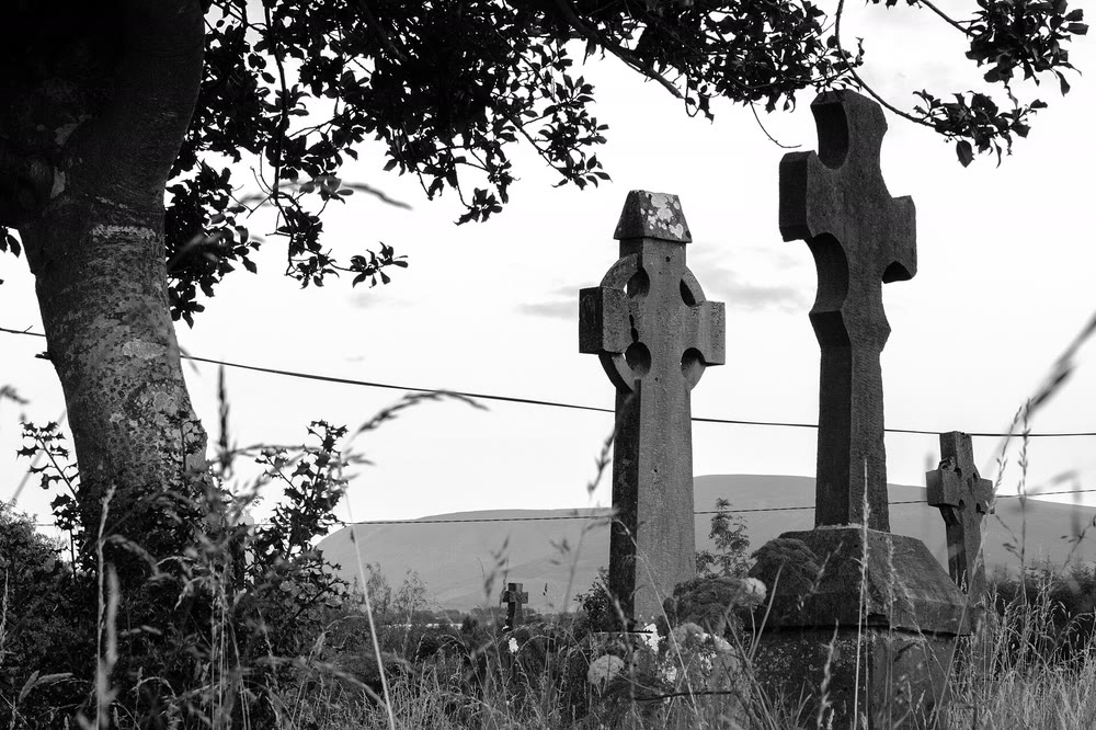 Black and white photo of tall Celtic cross headstones among long grass in a country graveyard, with a tree and a hill behind