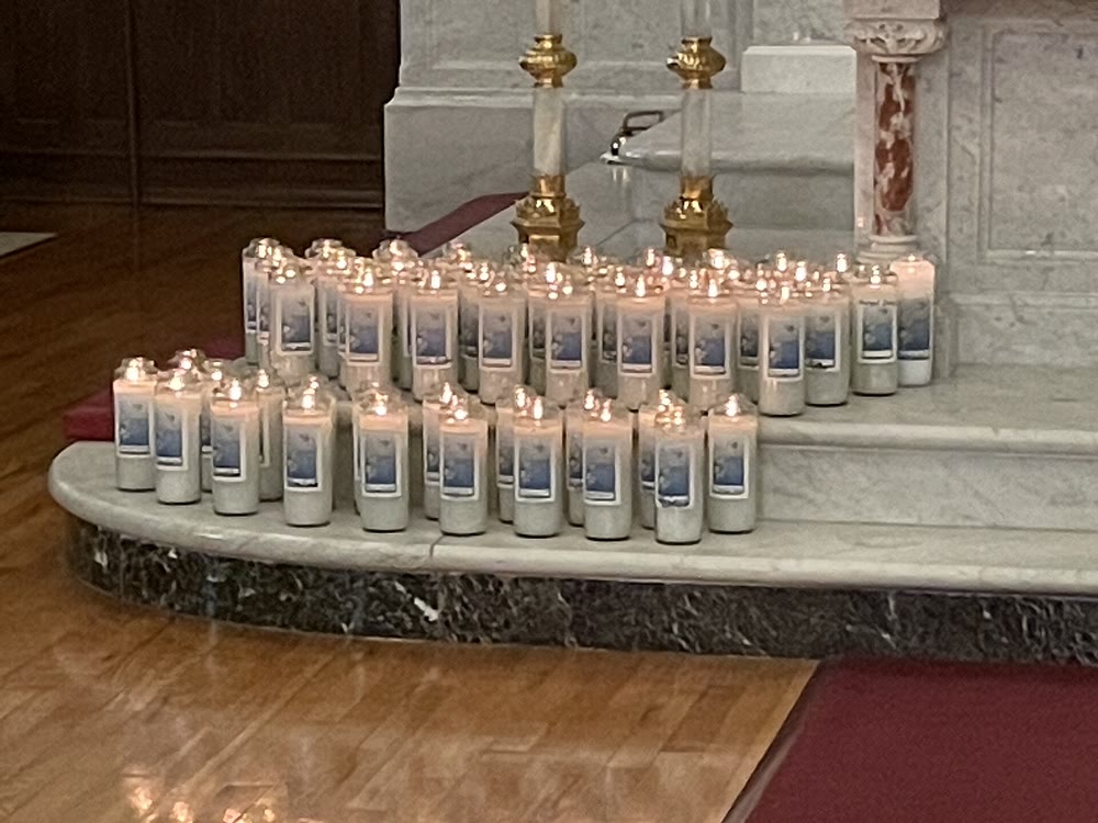 Rows of lit memorial candles in tall glass holders on the marble altar steps of a church