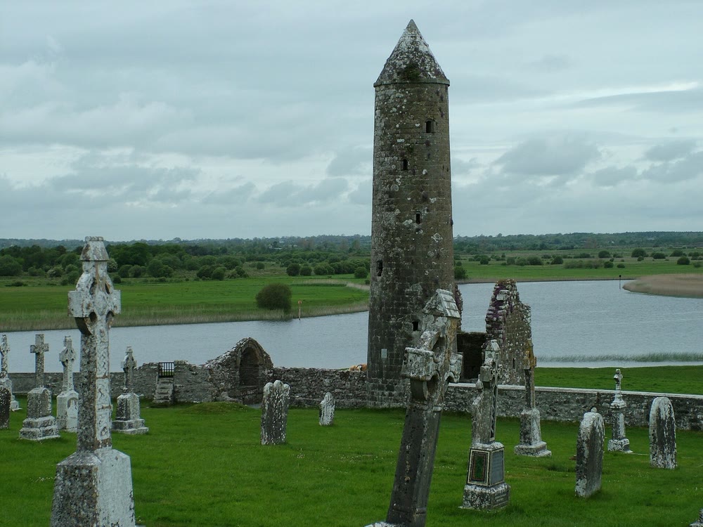 Round tower with a conical stone cap rising beside ruined church walls at Clonmacnoise, with old grave crosses in the grass and the River Shannon behind