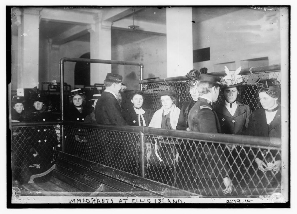 Black-and-white photograph of young women immigrants in hats and coats standing behind wire-mesh railings at Ellis Island as two uniformed officials talk with them