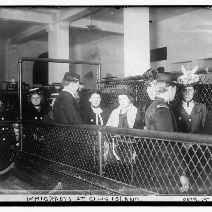 Black-and-white photograph of young women immigrants in hats and coats standing behind wire-mesh railings at Ellis Island as two uniformed officials talk with them