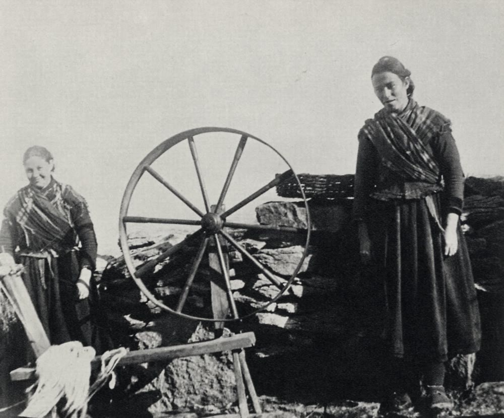 Black and white photograph of two Aran Island women in plaid shawls and long dark skirts beside a large wooden spinning wheel in front of a dry stone wall