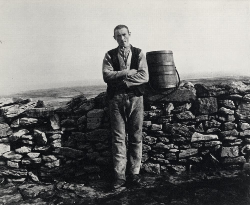 Black and white photograph of a man on Inis Meáin in a dark waistcoat over a pale jacket, arms folded, standing against a dry stone wall beside a small wooden barrel
