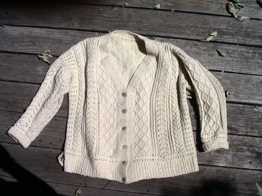 Cream Aran cardigan with buttons, laid flat on weathered wooden boards, showing vertical panels of cables, diamonds and openwork stitches