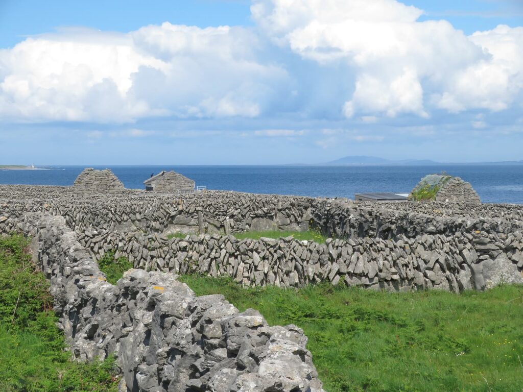 Dry stone walls criss-crossing green fields on Inis Me&aacute;in, with small stone barns and the sea beyond under a cloudy blue sky