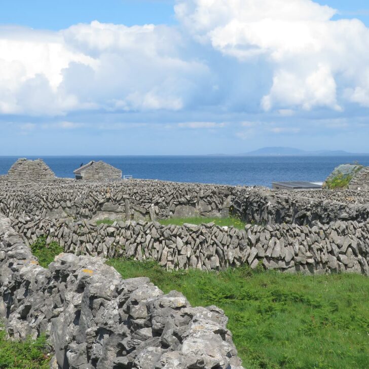 Dry stone walls criss-crossing green fields on Inis Meáin, with small stone barns and the sea beyond under a cloudy blue sky