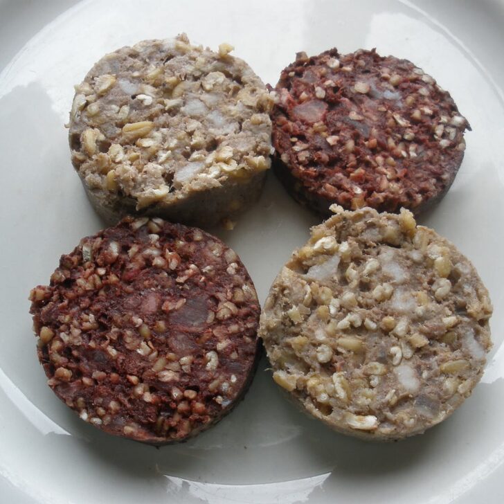 Four uncooked round slices of Irish pudding on a white plate: two dark red-brown black pudding slices and two pale white pudding slices, all studded with oatmeal
