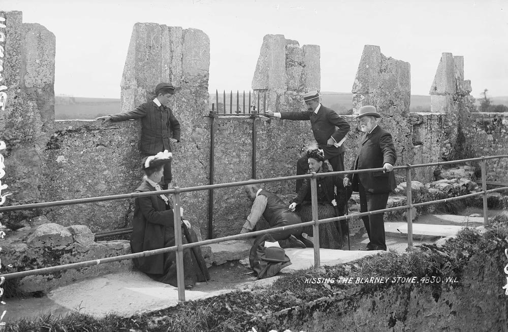 Black-and-white photo from about 1897 of Blarney Castle's battlements: Victorian visitors in hats and long coats stand and sit beside an iron grille in the parapet, while one man crouches at the edge of the walkway