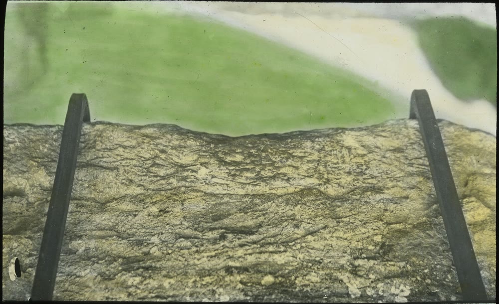 Hand-coloured 1913 lantern slide looking down at the Blarney Stone: a long block of weathered grey stone with two curved iron handles, and green lawn far below