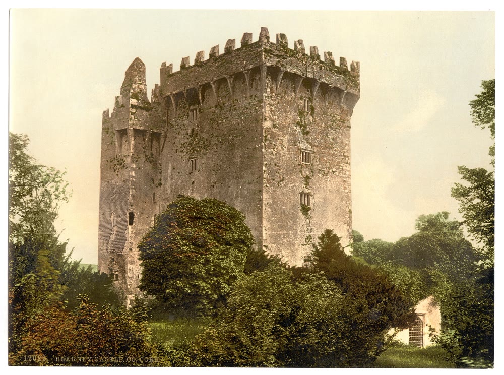 Colour-tinted 1890s print of Blarney Castle's tall, square stone keep with jagged battlements and overhanging parapet, rising above thick trees and shrubs