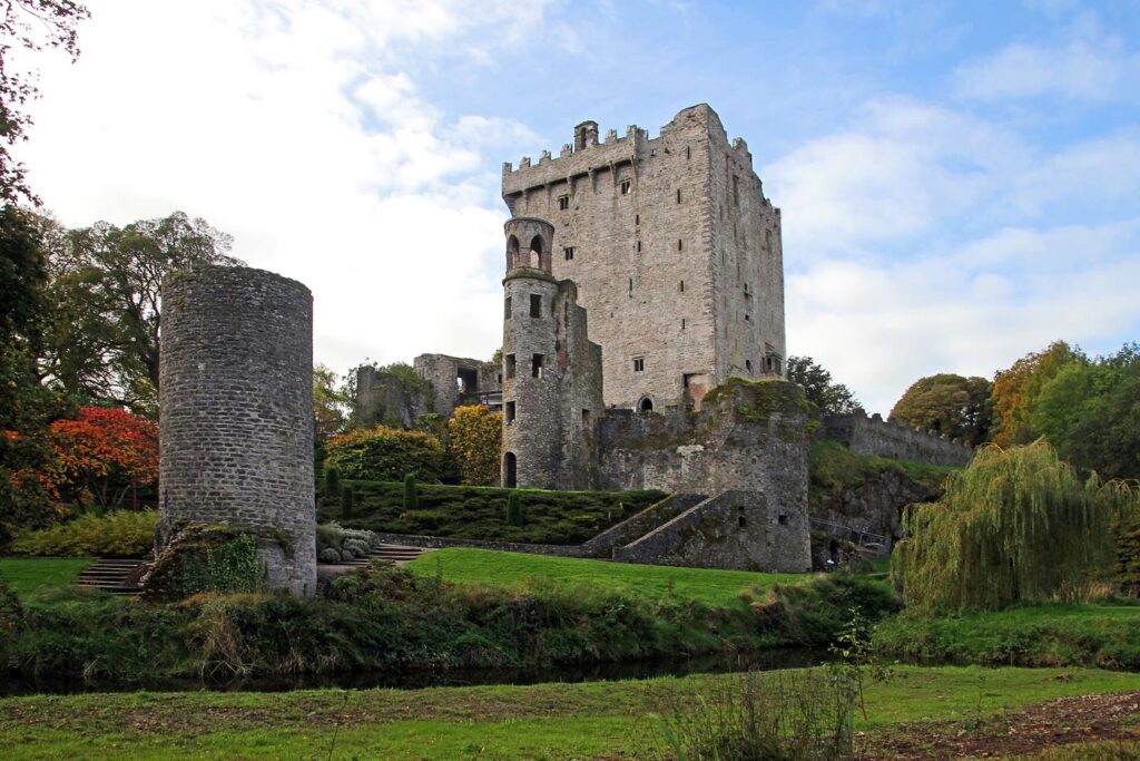 Blarney Castle's tall grey stone keep and round turret rising above green lawns, a stream and autumn trees, with a ruined round tower on the left