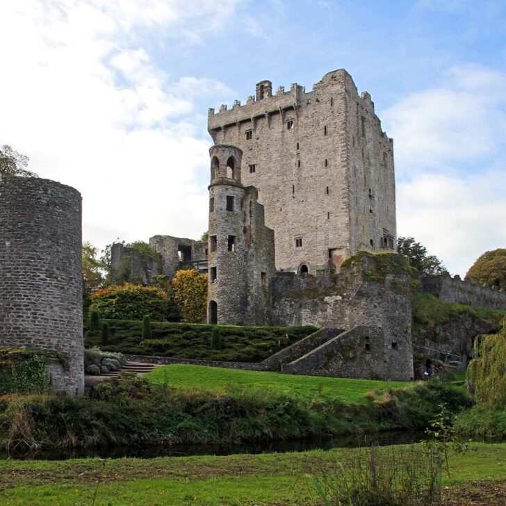 Blarney Castle's tall grey stone keep and round turret rising above green lawns, a stream and autumn trees, with a ruined round tower on the left
