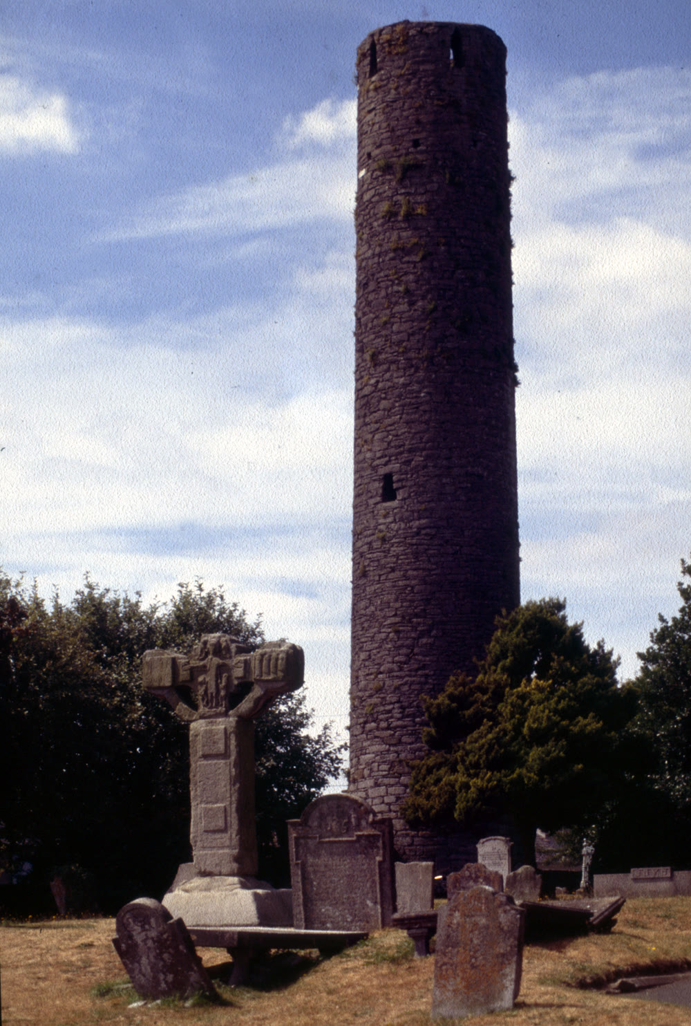 Tall stone round tower beside a weathered stone high cross and old gravestones in the churchyard at Kells, Co. Meath