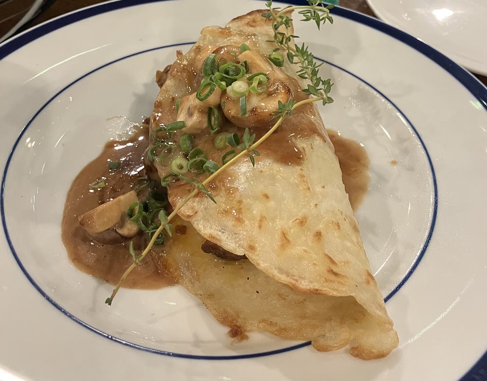 A thin pan boxty folded over a savoury filling in a brown mushroom sauce, topped with sliced scallions and a sprig of thyme, on a white plate with a blue rim