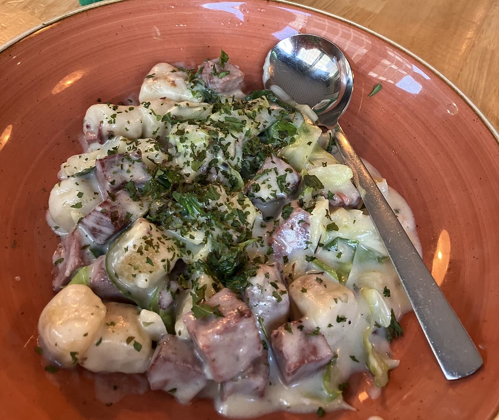 A terracotta bowl of boxty dumplings with chunks of corned beef and cabbage in a creamy sauce flecked with parsley, with a spoon resting in the bowl