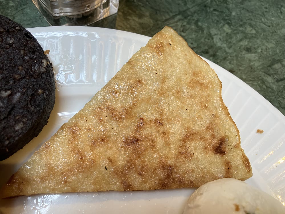 A golden, pan-fried triangle of boxty on a white plate, next to a round slice of black pudding