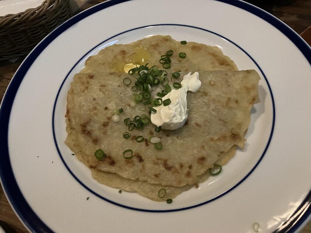 Two thin, golden pan boxty pancakes on a blue-rimmed white plate, topped with a spoonful of sour cream, melting butter and sliced scallions