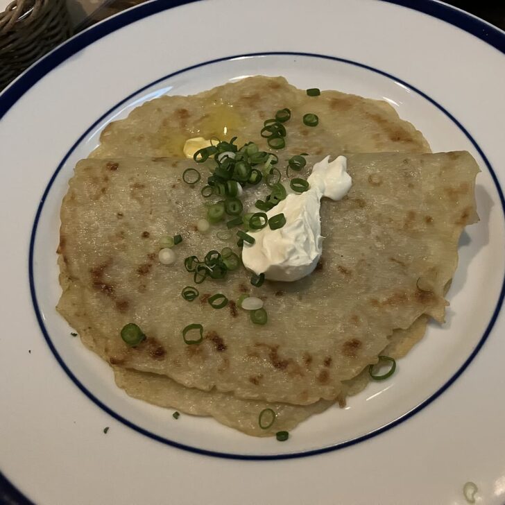 Two thin, golden pan boxty pancakes on a blue-rimmed white plate, topped with a spoonful of sour cream, melting butter and sliced scallions