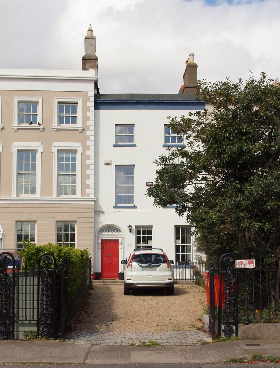 A white three-storey Georgian terraced house with a red front door and fanlight, a gravel drive and black iron railings at 15 Marino Crescent, Clontarf