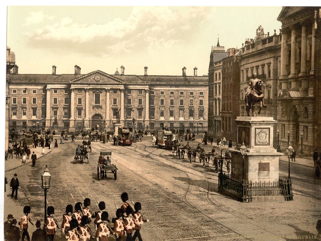 Colour photochrom of College Green, Dublin, in the 1890s: the pillared front of Trinity College behind horse trams and carriages, with an equestrian statue on a tall plinth to the right and soldiers marching in the foreground