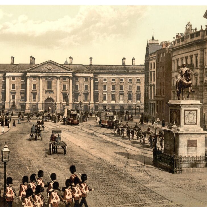Colour photochrom of College Green, Dublin, in the 1890s: the pillared front of Trinity College behind horse trams and carriages, with an equestrian statue on a tall plinth to the right and soldiers marching in the foreground
