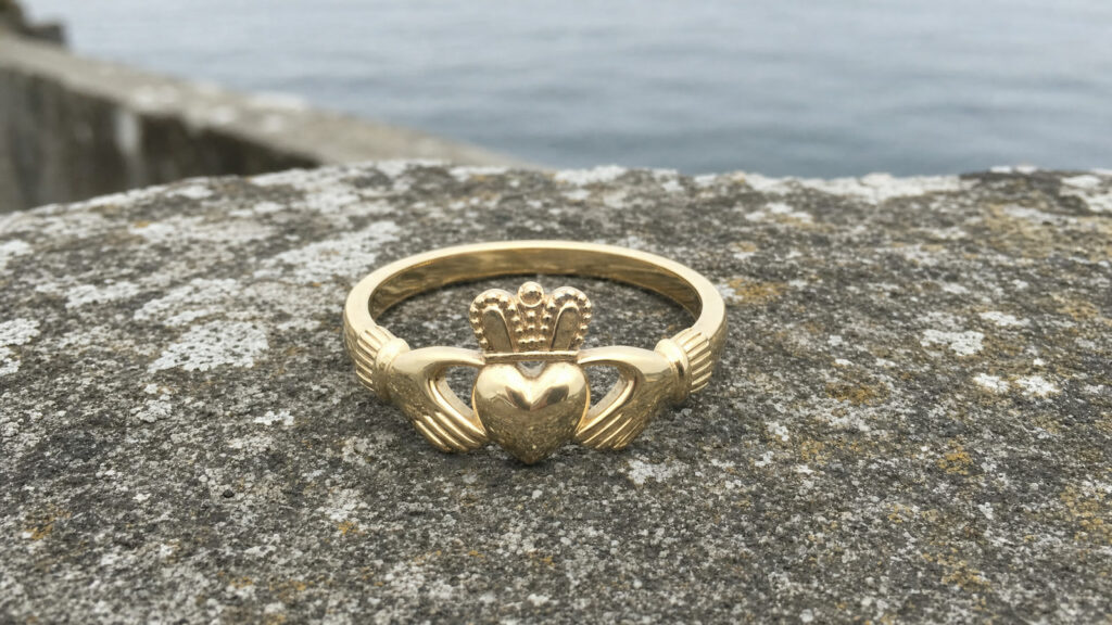 A gold Claddagh ring with two hands holding a crowned heart, resting on a stone wall by the sea