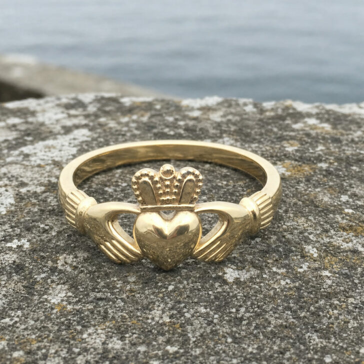 A gold Claddagh ring with two hands holding a crowned heart, resting on a stone wall by the sea
