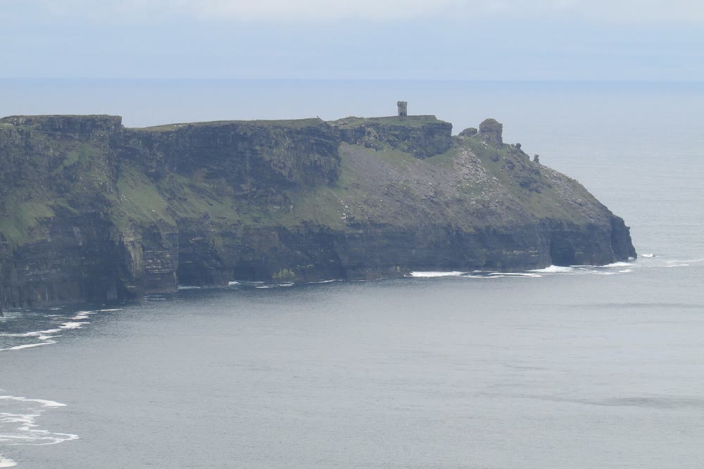 A long dark headland jutting into a calm grey sea, with a small square stone tower on its flat green top