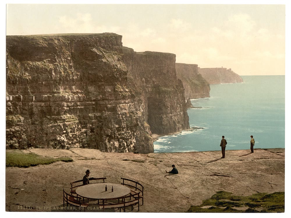 Tinted 1890s photograph of tall layered cliffs above a turquoise sea, with men in hats on a flat stone platform and a round table with curved iron seats in the foreground