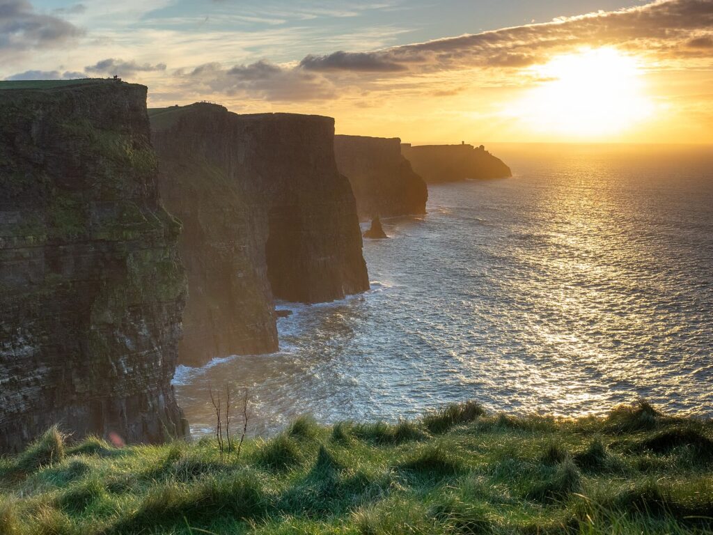 The Cliffs of Moher at sunset: dark layered cliffs step away to a distant headland with a small tower, a sea stack stands below and the low sun lights the sea, with grass in the foreground