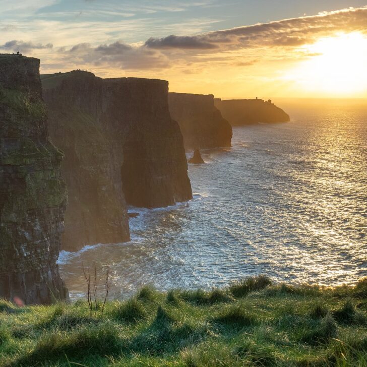 The Cliffs of Moher at sunset: dark layered cliffs step away to a distant headland with a small tower, a sea stack stands below and the low sun lights the sea, with grass in the foreground
