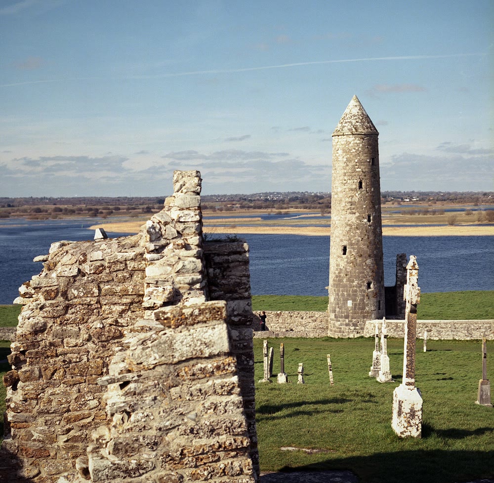 McCarthy's Tower at Clonmacnoise: a round stone tower with a conical cap beside a ruined church wall, with grave crosses in the grass and the wide River Shannon behind