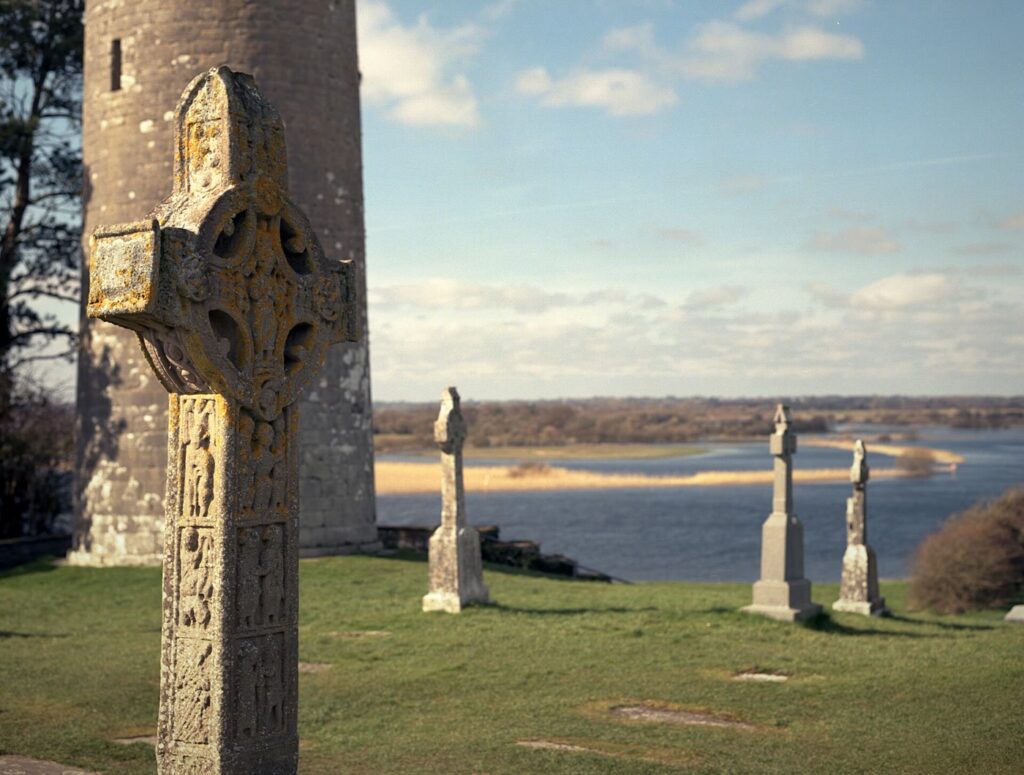 Clonmacnoise: a lichen-covered replica high cross in front of O'Rourke's round tower, with grave crosses on the grass and the wide River Shannon and its flood meadows beyond