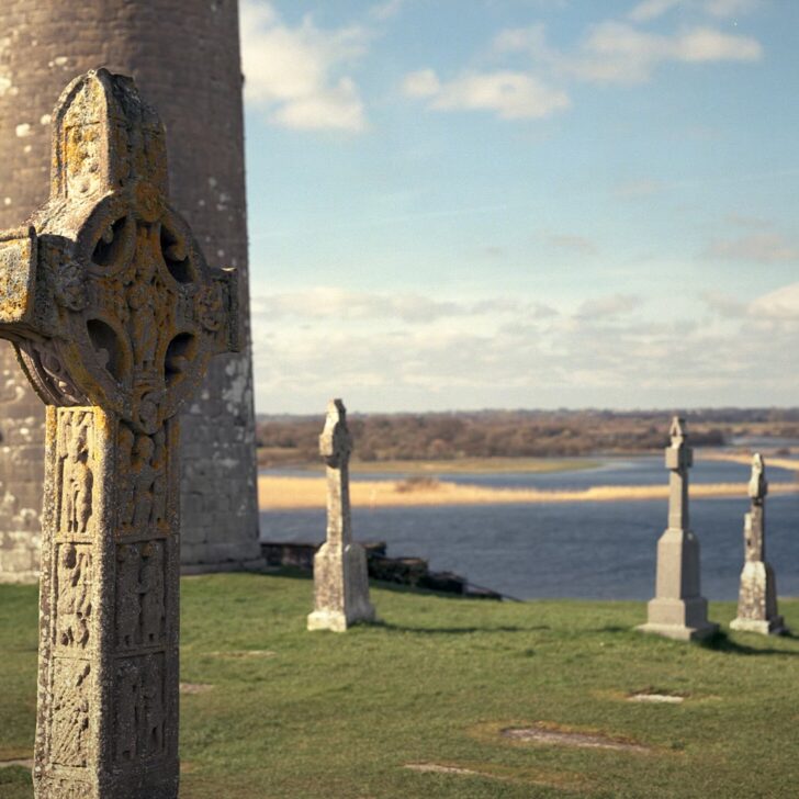 Clonmacnoise: a lichen-covered replica high cross in front of O'Rourke's round tower, with grave crosses on the grass and the wide River Shannon and its flood meadows beyond