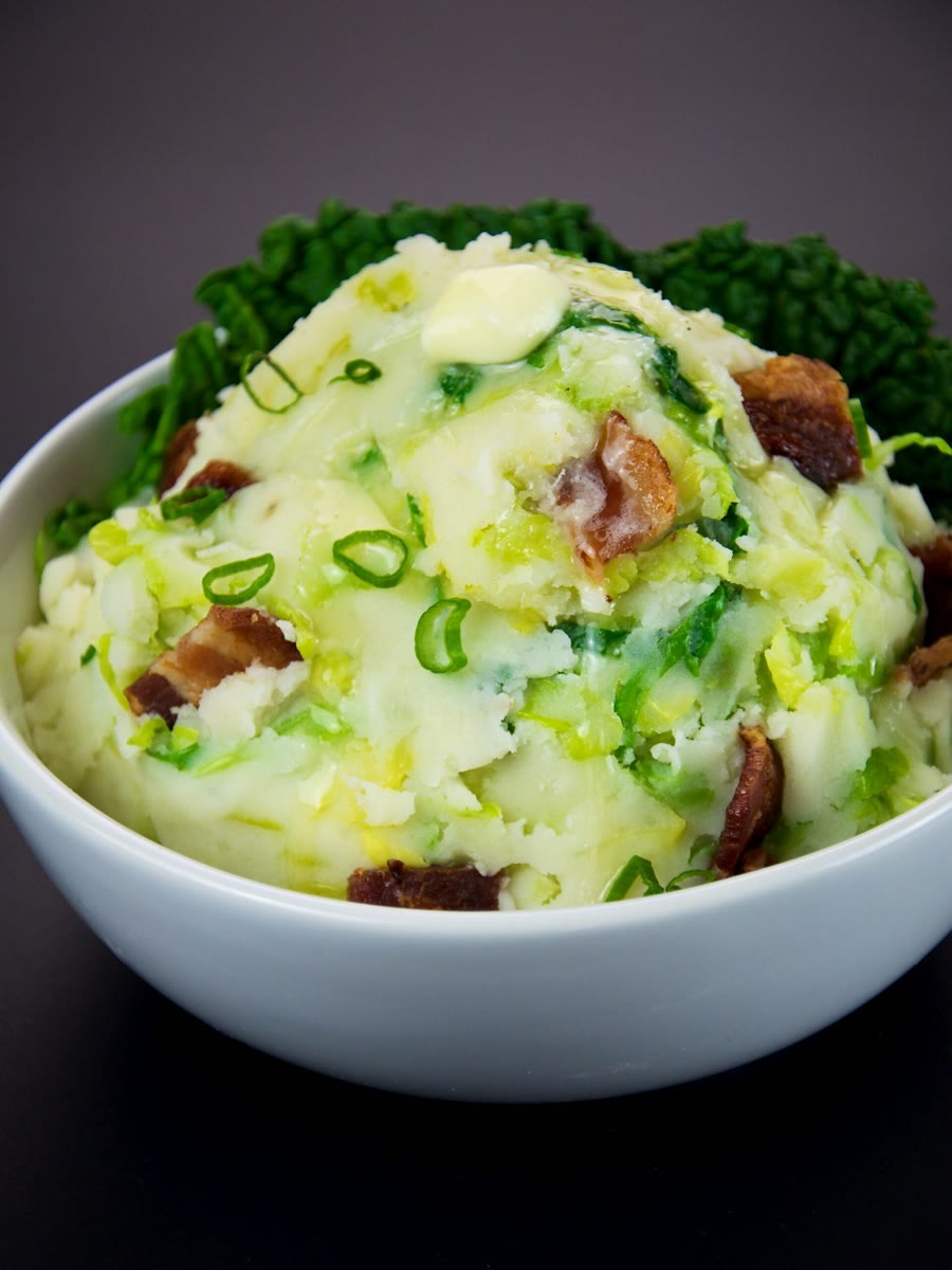 A white bowl of colcannon flecked with Savoy cabbage, sliced scallions and crisp bacon, topped with a pat of melting butter, with a Savoy cabbage leaf behind