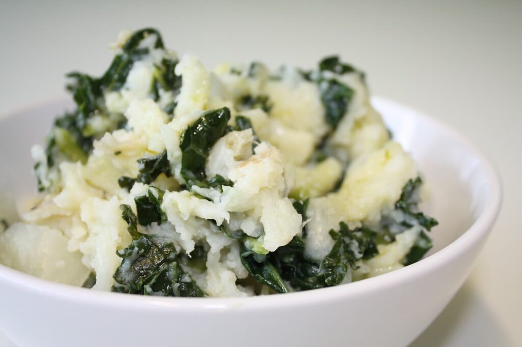 A white bowl of colcannon: fluffy mashed potato flecked with dark green kale, on a pale background