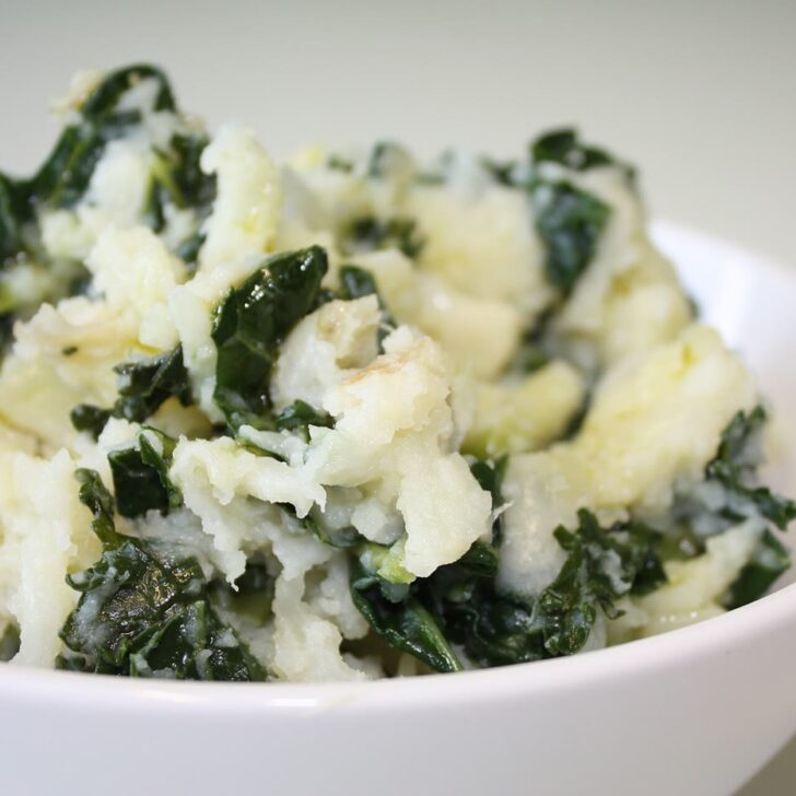 A white bowl of colcannon: fluffy mashed potato flecked with dark green kale, on a pale background
