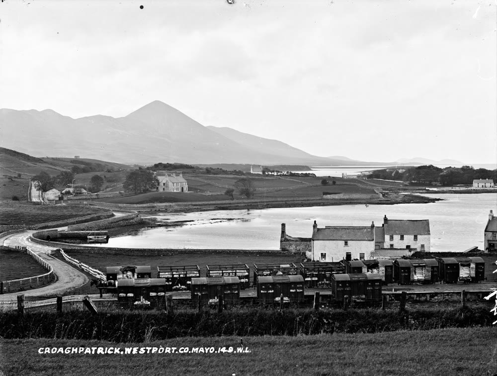 Old black and white photograph of the cone of Croagh Patrick rising beyond an inlet of Clew Bay, with whitewashed houses and a row of railway wagons near Westport in the foreground