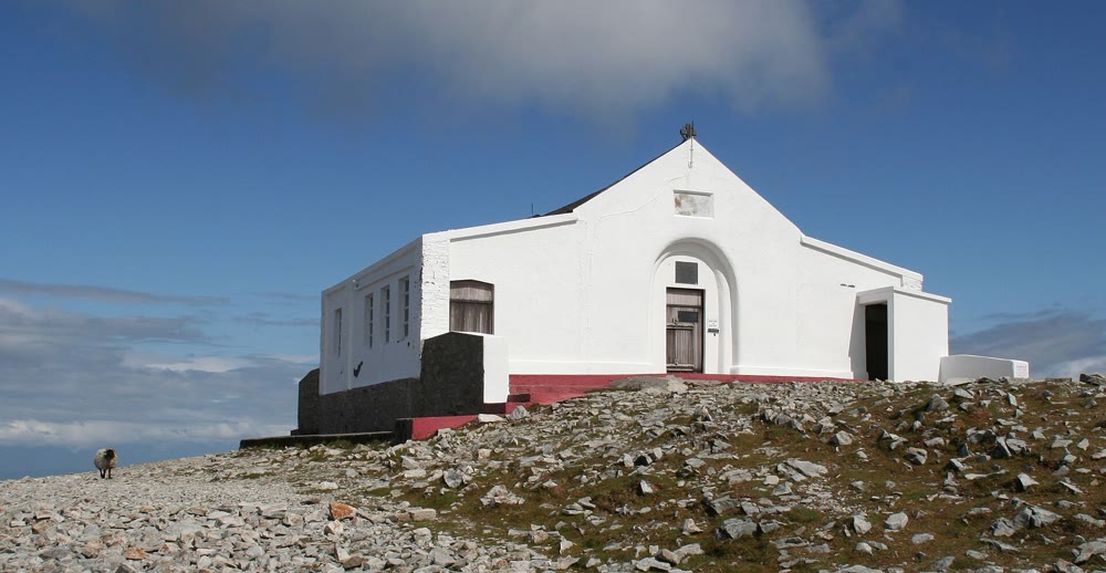 Small whitewashed chapel with a gabled front and an arched doorway on the stony summit of Croagh Patrick under a blue sky, with one sheep nearby