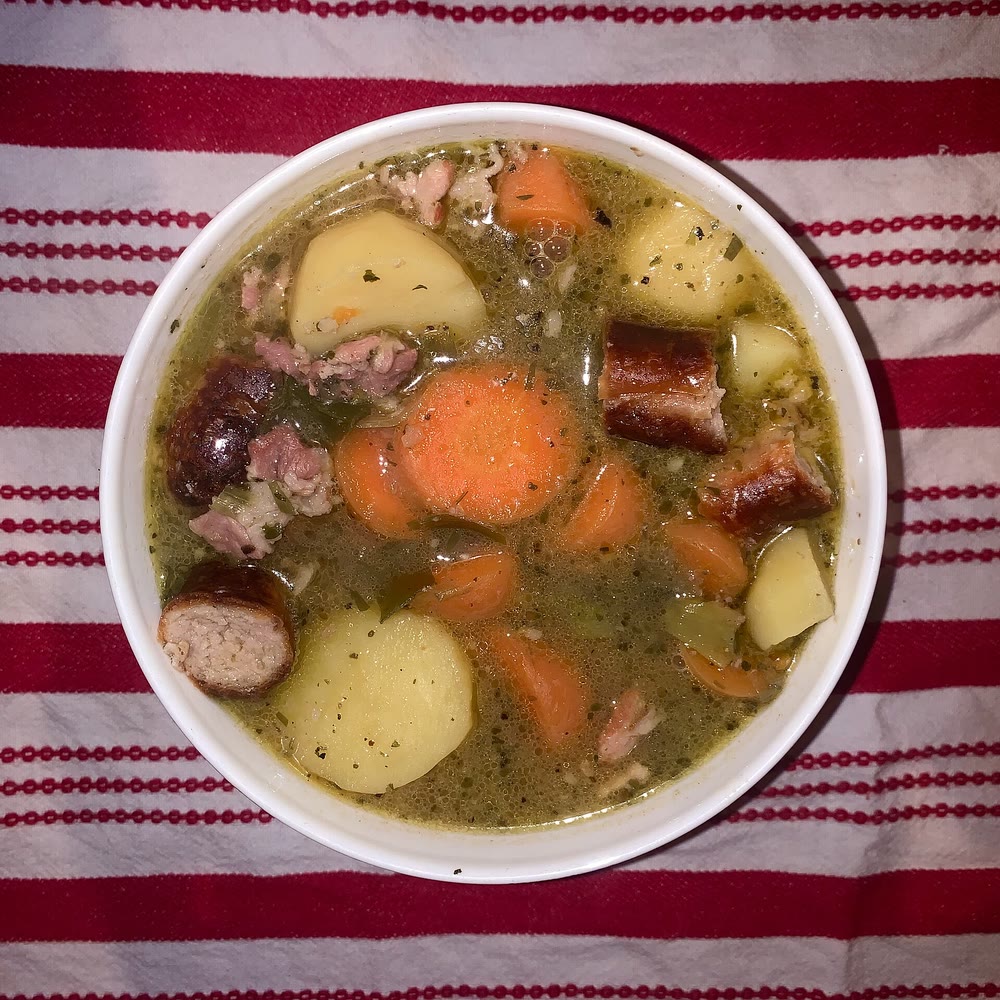 A white bowl of coddle seen from above, with browned sausage pieces, chunks of potato, sliced carrots and bacon in a herby broth, on a red and white striped cloth