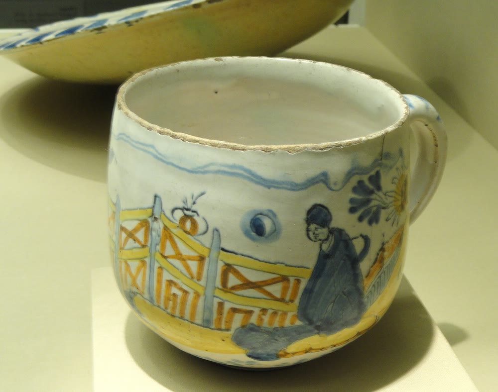 A small rounded cup with a handle, painted in blue, yellow and orange with a fence, a flower and a seated figure, in a museum case
