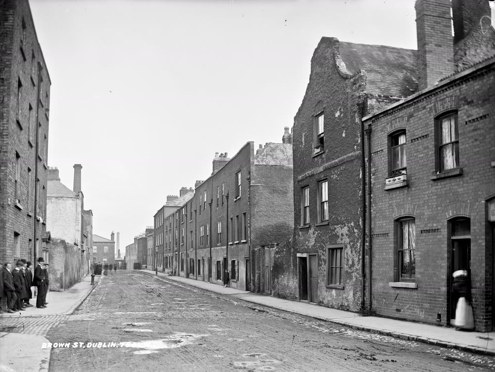 A black and white photo of a cobbled Dublin street lined with tall, worn brick houses, with a few people standing in doorways and on the footpath
