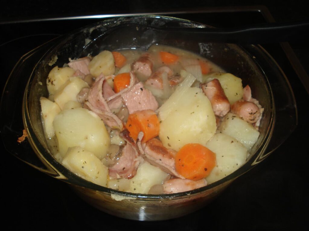 A glass casserole dish of oven-baked coddle: chunks of potato, sausage, bacon, carrot and onion in a pale broth flecked with dried herbs