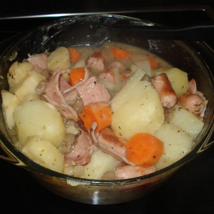 A glass casserole dish of oven-baked coddle: chunks of potato, sausage, bacon, carrot and onion in a pale broth flecked with dried herbs