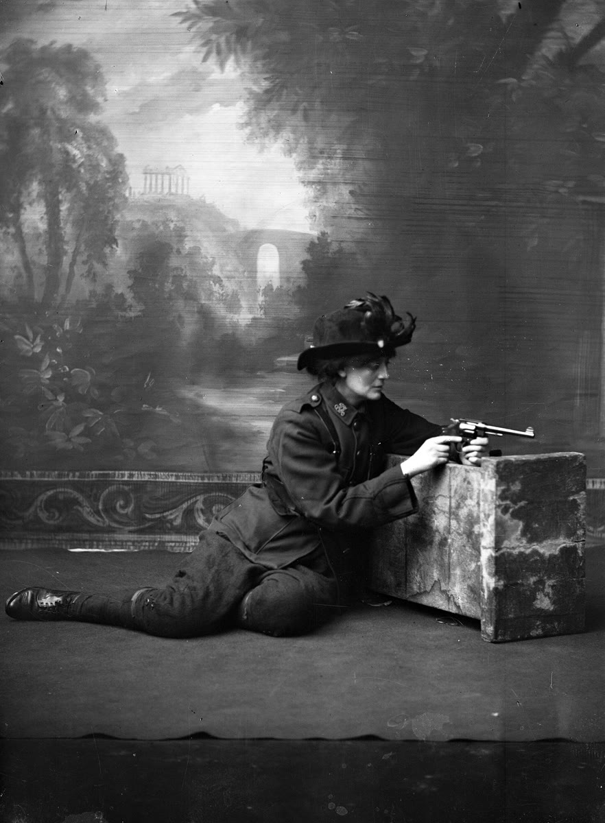 Studio portrait of Constance Markievicz in uniform and a plumed hat, sitting on the floor in front of a painted backdrop and aiming a revolver over a low block