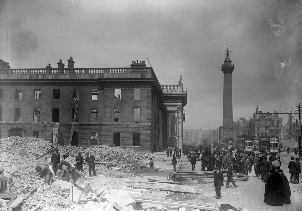 The roofless, burned-out shell of the GPO on Sackville Street in May 1916, with rubble and workmen in front, people and trams on the street and Nelson's Pillar rising behind