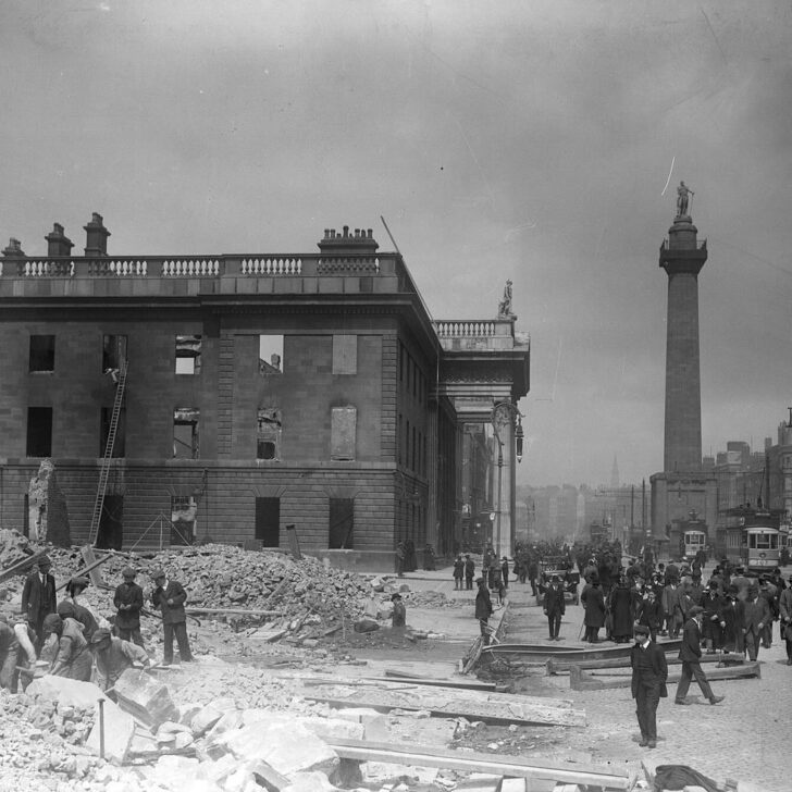 The roofless, burned-out shell of the GPO on Sackville Street in May 1916, with rubble and workmen in front, people and trams on the street and Nelson's Pillar rising behind
