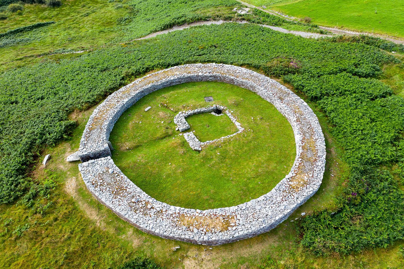 Aerial view of Knockdrum stone fort in Co. Cork: a circular dry-stone wall enclosing a grassy interior with the low stone outline of a rectangular building, surrounded by ferns