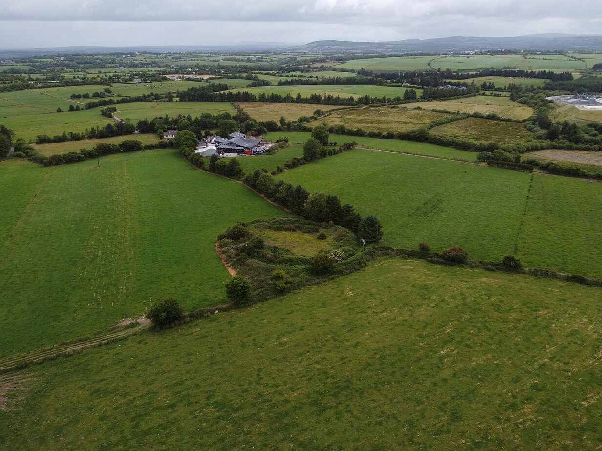 Aerial view of an overgrown circular enclosure ringed with bushes in a green field in Co. Cork, with farm buildings and patchwork fields stretching to hills behind